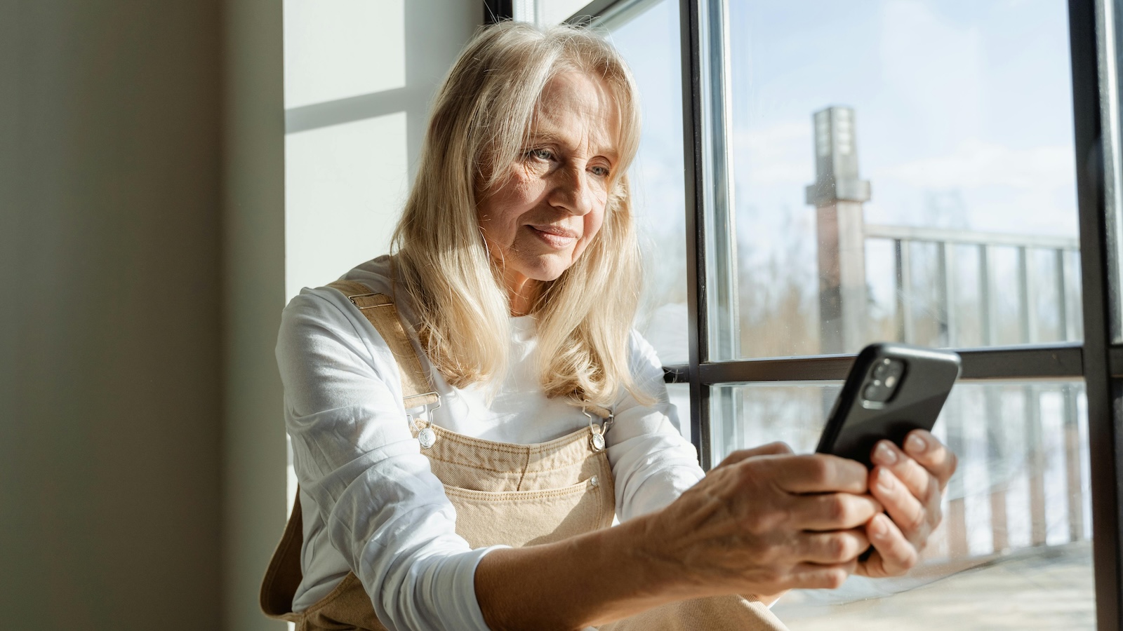 Older woman with white blonde hair in a white shirt holding a phone and sitting against a big window 